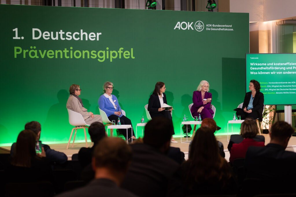 Vier Frauen sitzen auf einer Bühne vor grünem Hintergrund, auf dem steht Deutscher Präventionsgipfel AOK